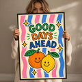 A woman holding a framed poster with a colorful design featuring two smiling fruits, an orange and a lemon, along with the text "GOOD DAYS AHEAD".