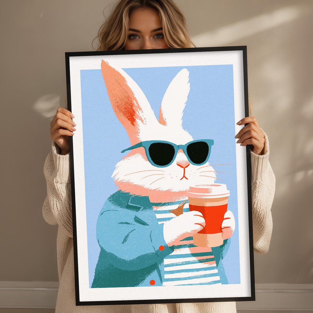 A woman is holding up a framed illustration of a white rabbit wearing sunglasses and holding a coffee cup.