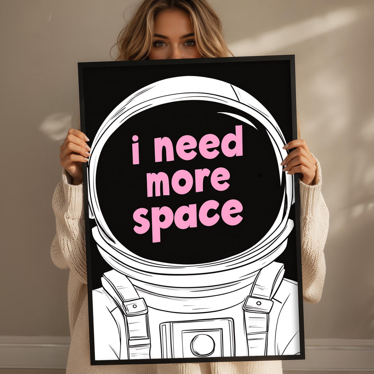 A woman holding a framed poster with a space helmet and the text "I need more space" written on it.
