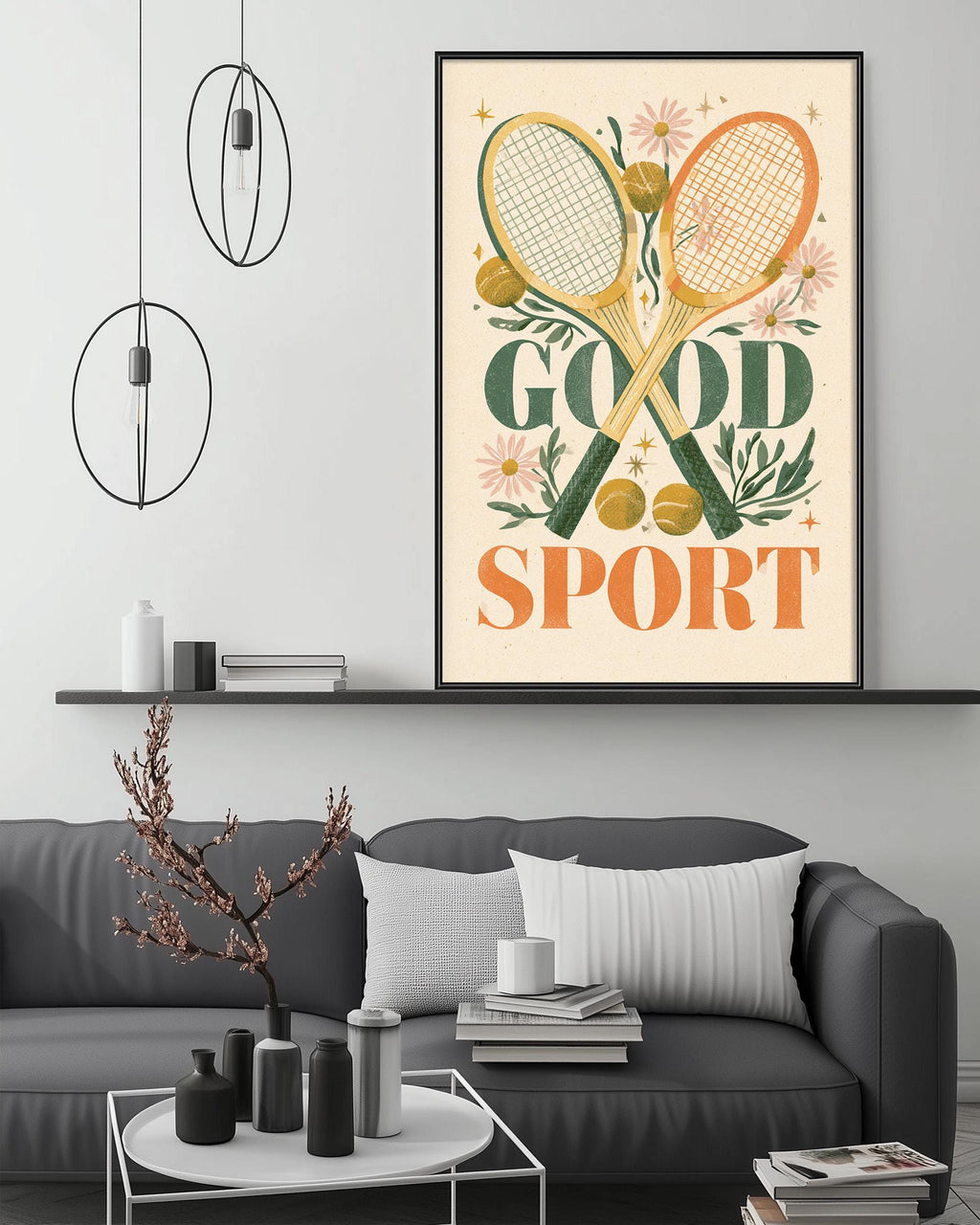 a living room with a gray couch, a coffee table, and a framed poster on the wall. The poster has a tennis racket and flowers as its main design elements, with the text "GOOD SPORT" prominently displayed.