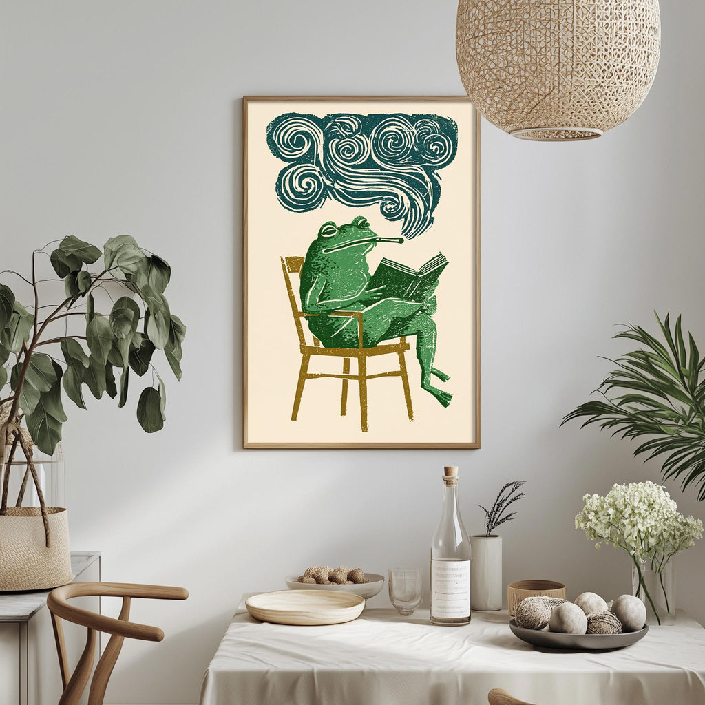 a framed illustration of a frog sitting in a chair, reading a book, with a cloud-like design above it. The setting includes a dining table with a bowl of fruit, a bottle, and a vase with flowers.