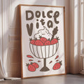 A framed poster with a drawing of a bowl of strawberries and the text "Dolce Vita" on it, placed on a wooden floor next to a window.