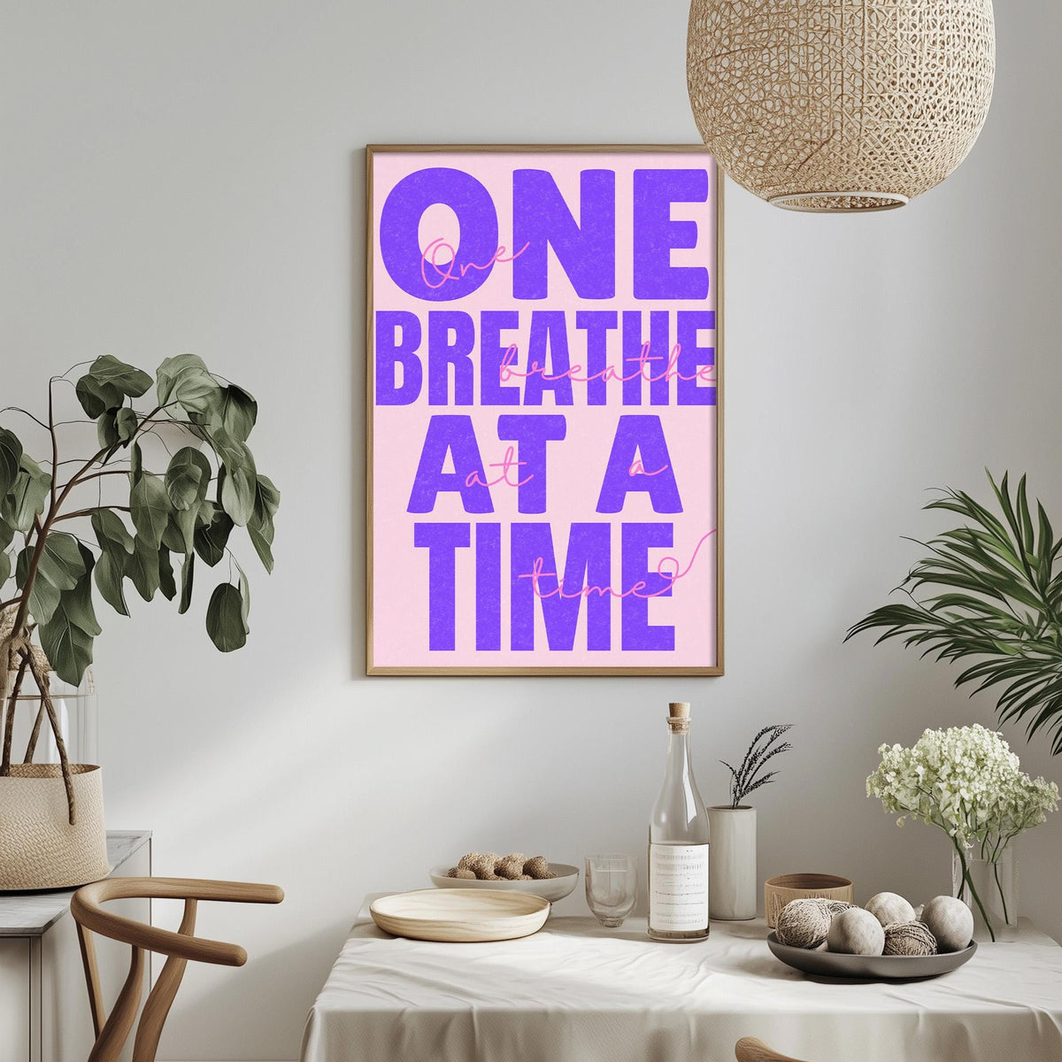 a framed poster with the text "ONE BREATHE AT A TIME" in purple and pink colors, hanging on a wall above a dining table with various items on it.