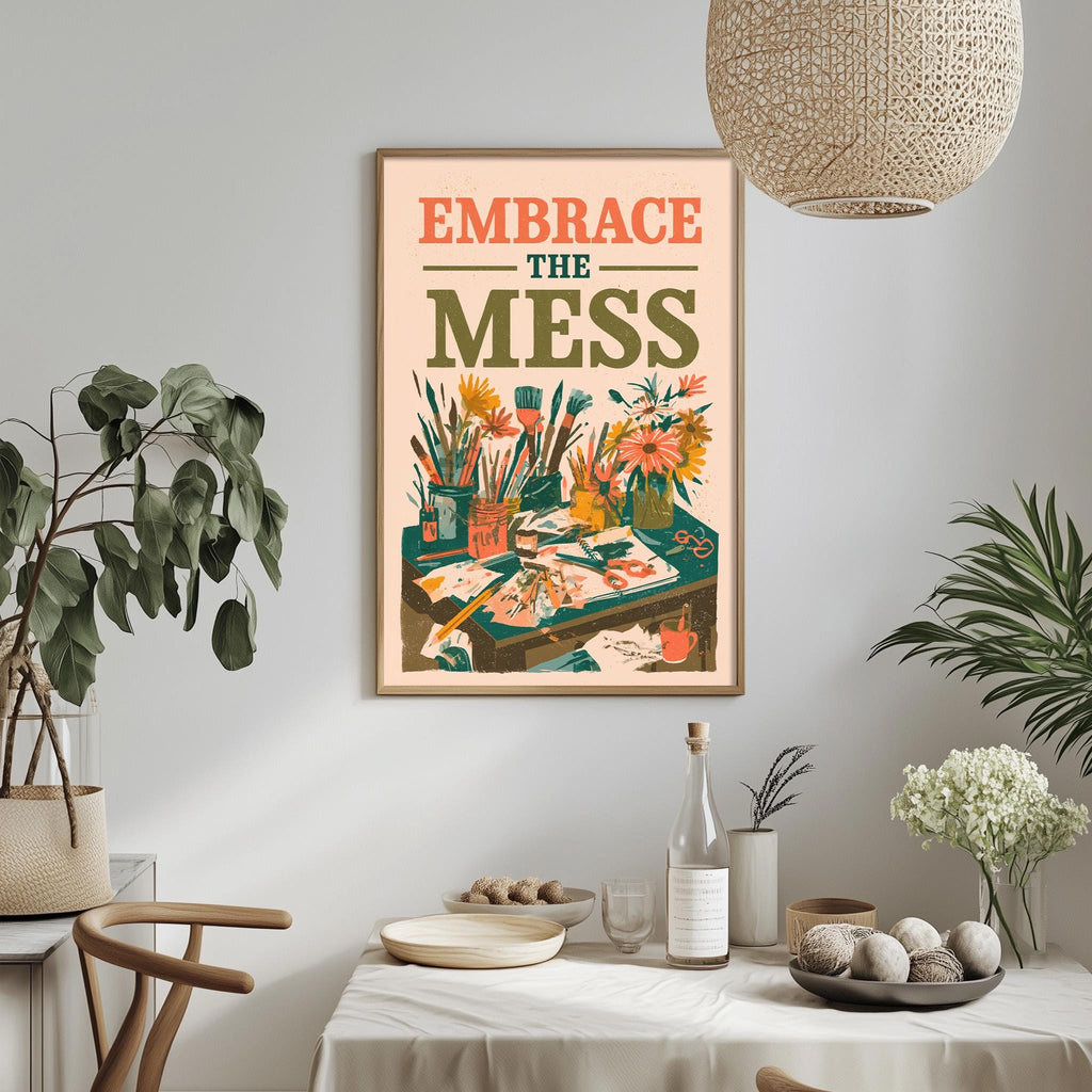 a framed poster with the text "Embrace the mess" hanging on a wall above a dining table with various items on it, including a bottle, bowls, and a vase.