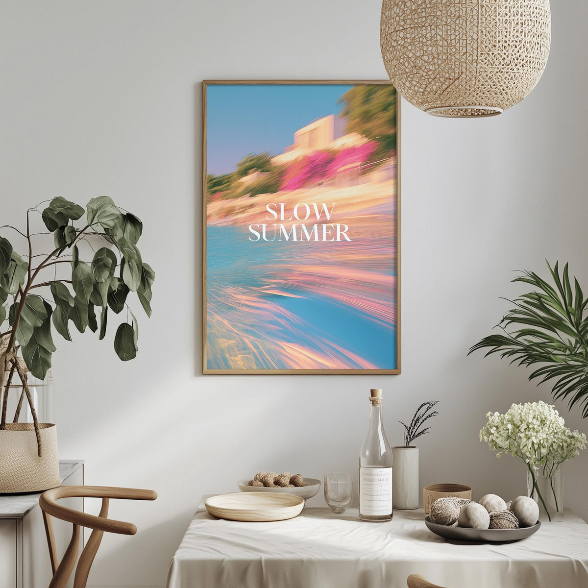 a dining table with a bowl of fruit, a bottle, and a vase of flowers, along with a potted plant and a hanging light fixture. Above the table, there is a framed poster with the text "SLOW SUMMER" displayed on it.