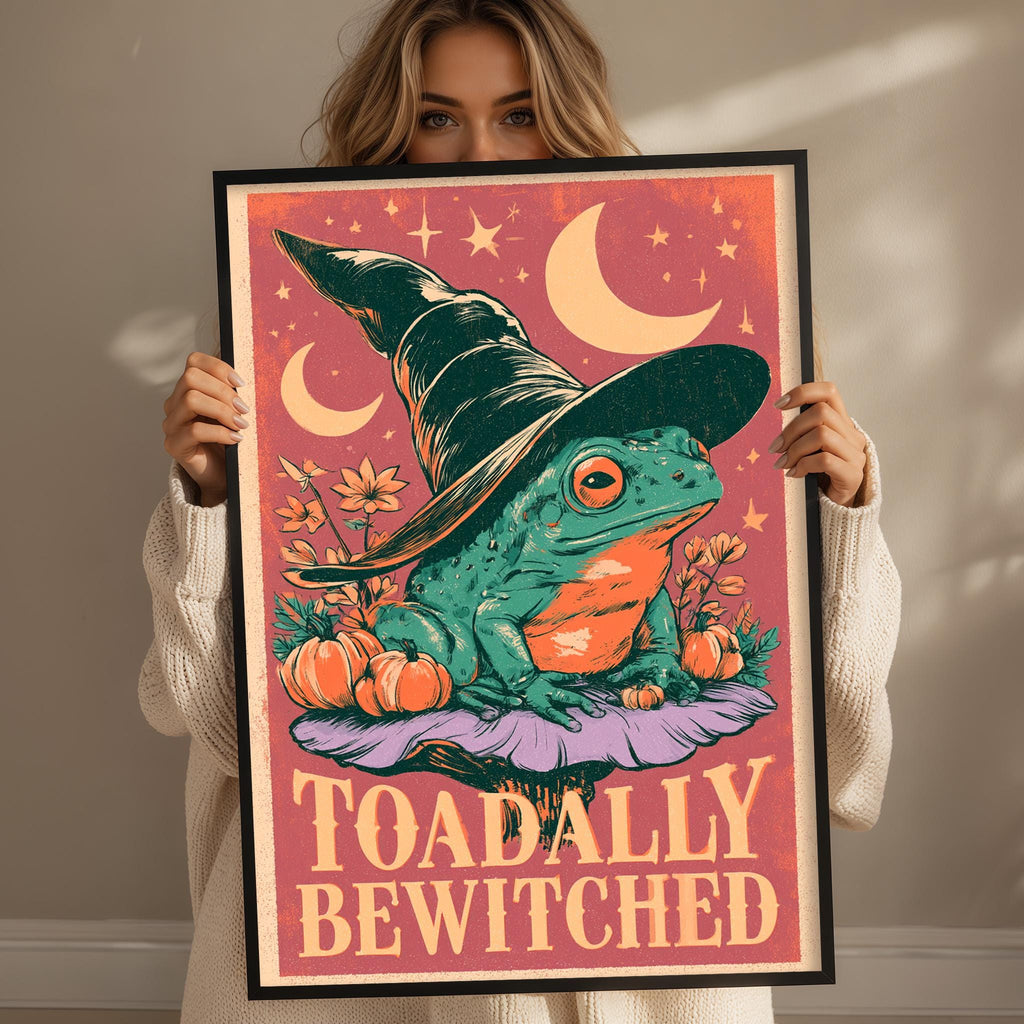 A woman is holding up a framed poster with a whimsical illustration of a frog wearing a witch's hat and surrounded by pumpkins and flowers.