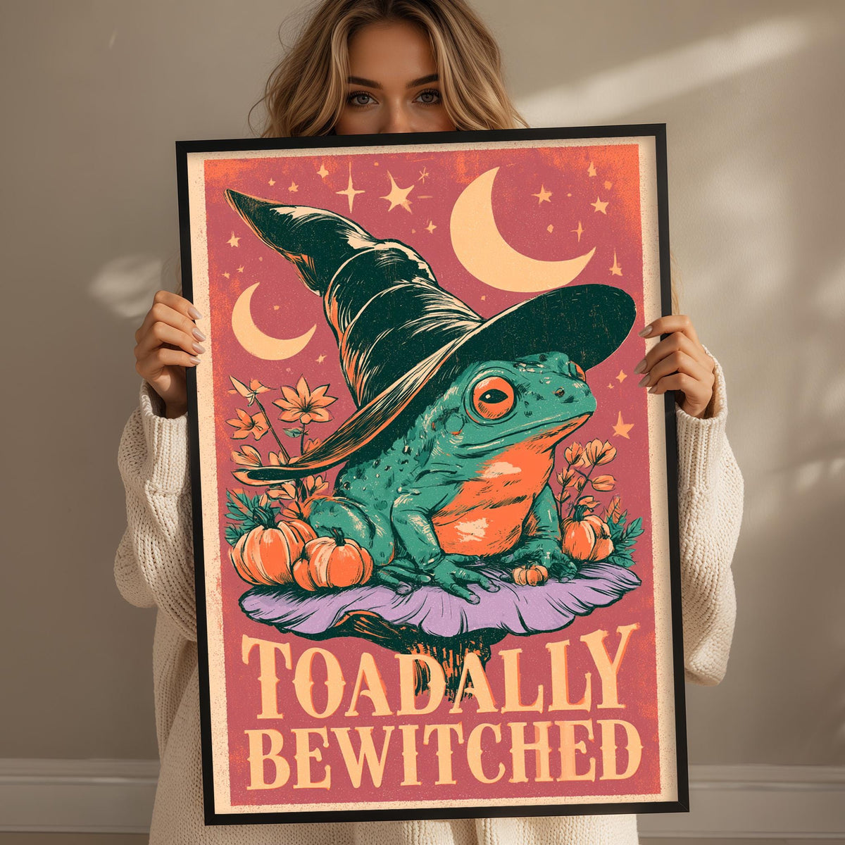A woman is holding up a framed poster with a whimsical illustration of a frog wearing a witch's hat and surrounded by pumpkins and flowers.