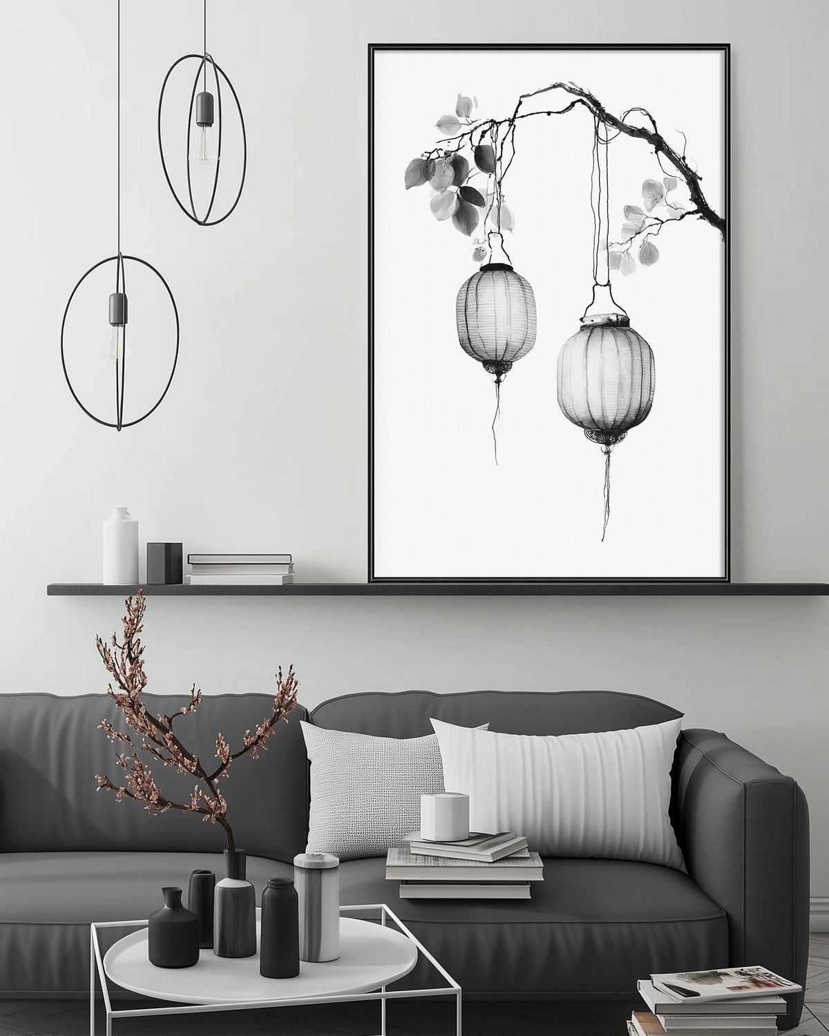 a modern living room with a gray sofa, a coffee table, and a large framed artwork featuring two hanging lanterns on a branch. The room has a minimalist and elegant design, with a black and white color scheme and a few decorative elements such as a potted plant and a vase.