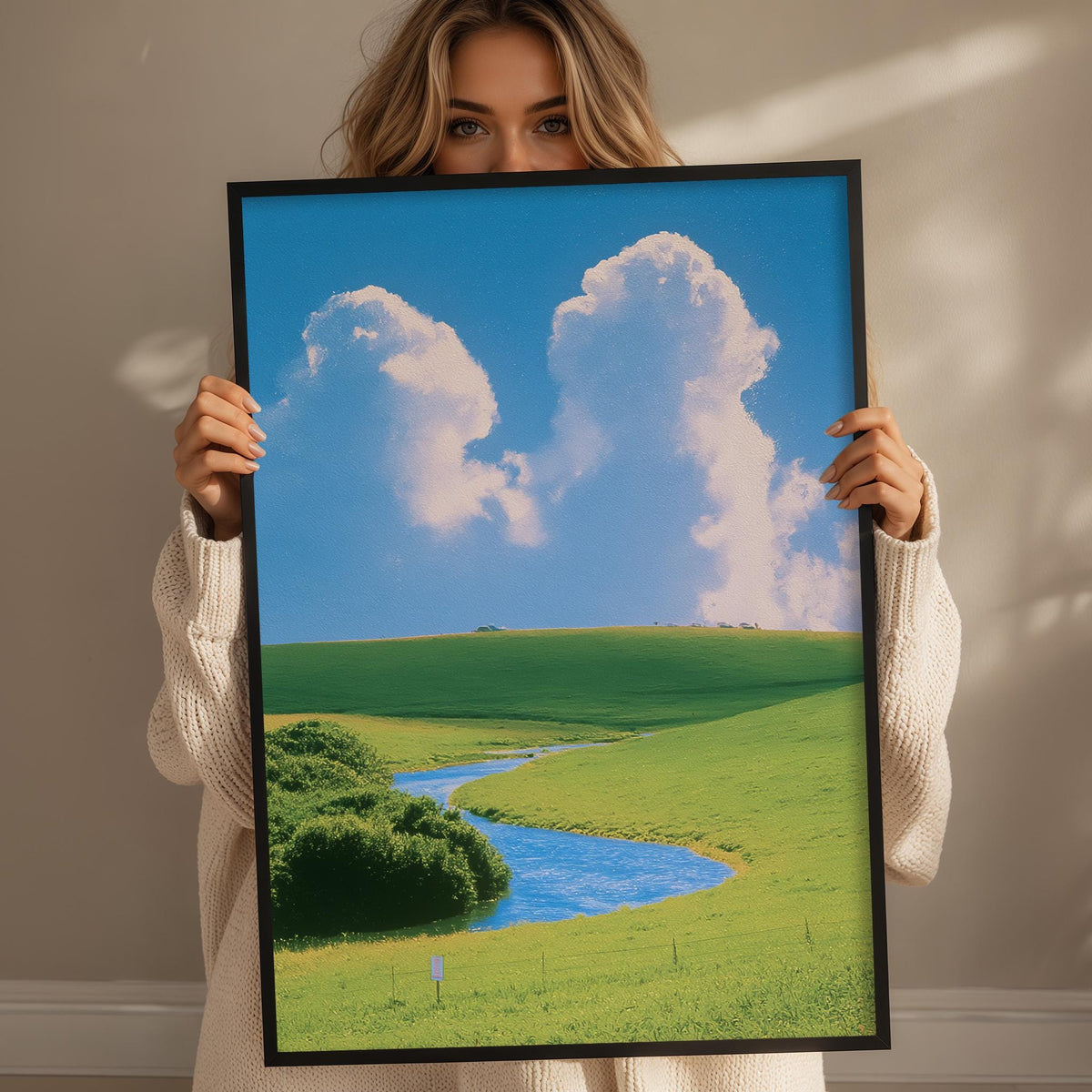 A woman holds up a framed picture of a serene landscape with a winding river, lush green fields, and a blue sky with fluffy white clouds.