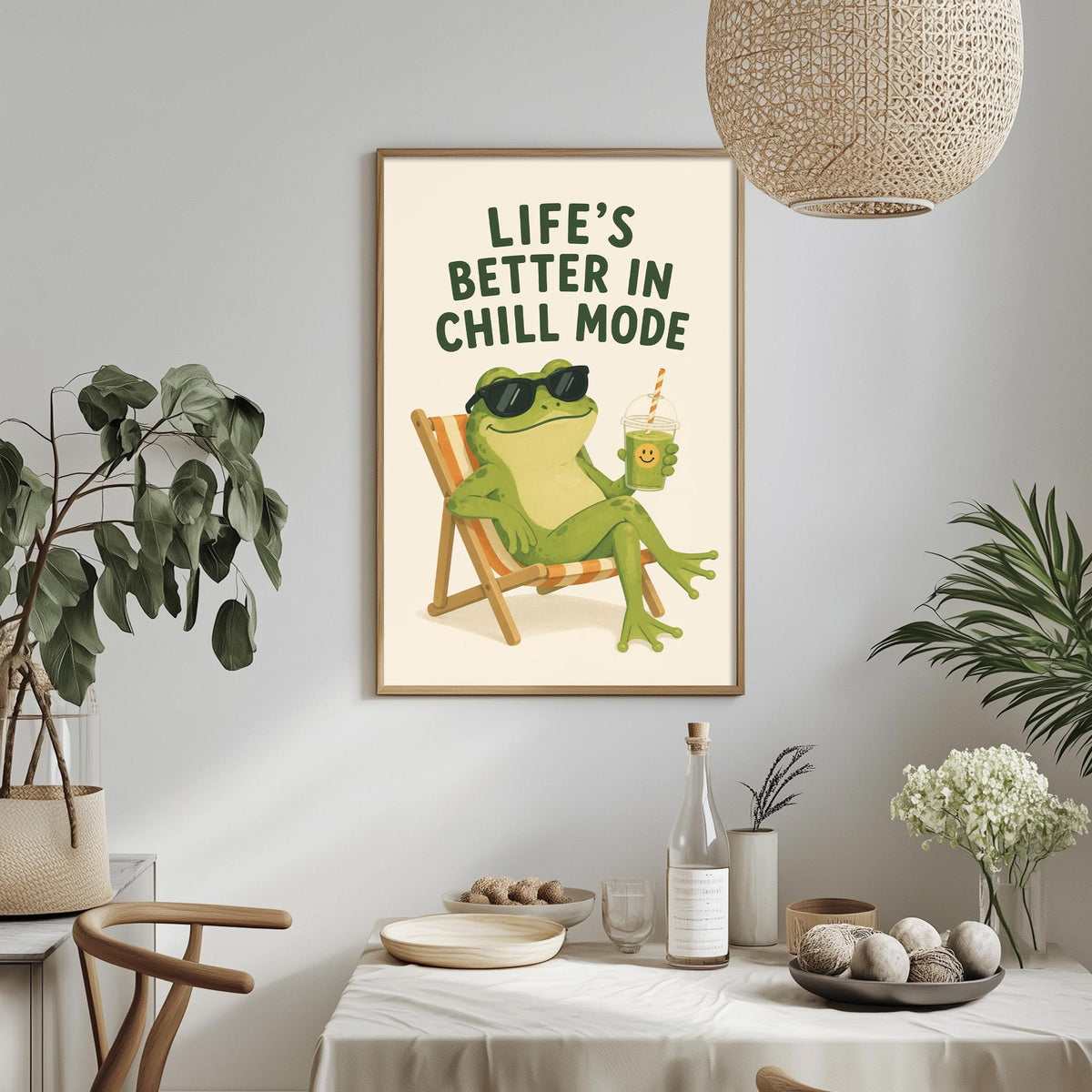 a framed poster with a cartoon frog sitting in a chair, holding a drink and wearing sunglasses, with the text "Life's better in chill mode" above it. The poster is hanging on a wall, and there is a dining table with a bowl, a bottle, and some plants in the foreground.