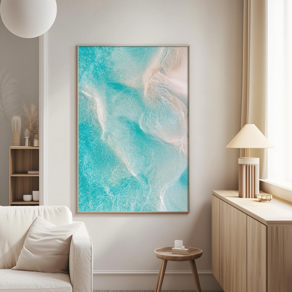 a living room with a large, vibrant painting of turquoise water hanging on the wall. The painting is framed in a wooden frame and is the focal point of the room. The living room also includes a white sofa, a wooden side table, and a lamp on a nearby table.