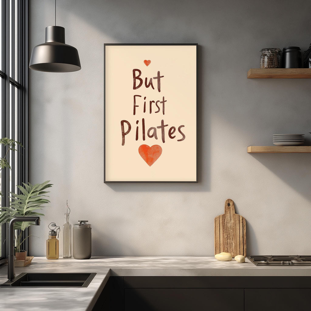 a kitchen counter with various kitchen items, such as a cutting board, a sink, and a potted plant. Above the counter, there is a framed poster with the text "But First Pilates" and a heart symbol.