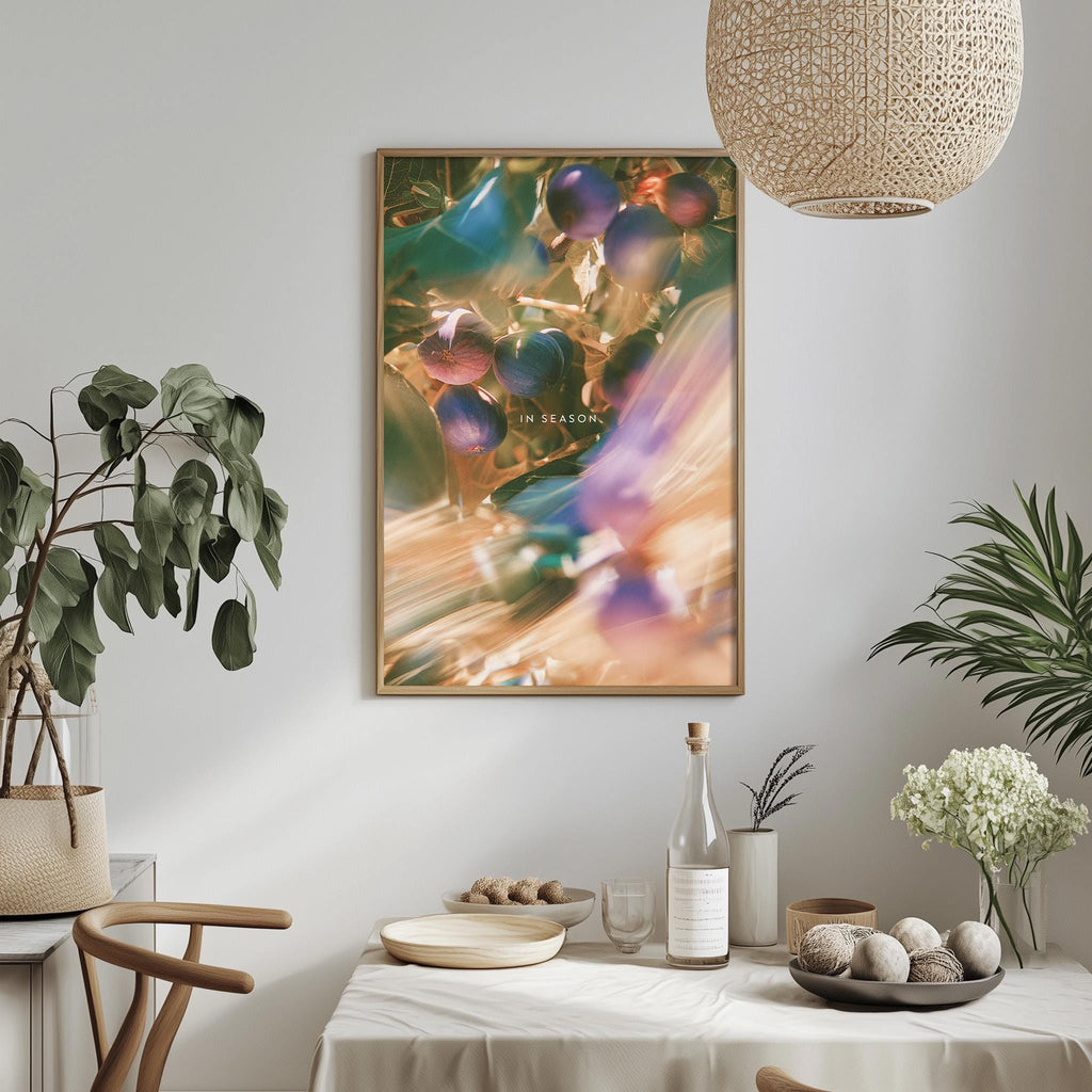a dining table with a white tablecloth, a bowl of fruit, a bottle, and a vase with flowers. A framed print of colorful fruits hangs on the wall above the table.