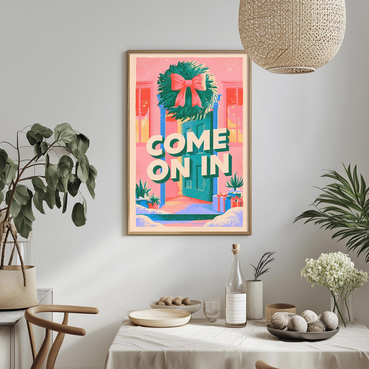 a dining table with a white tablecloth, a bowl of fruit, a bottle, and a vase with flowers. Above the table, there is a framed poster with the text "COME ON IN" in a colorful design.