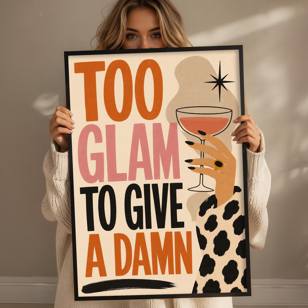 A woman holding a framed poster with the text "Too Glam to Give a Damn" and an illustration of a hand holding a wine glass.