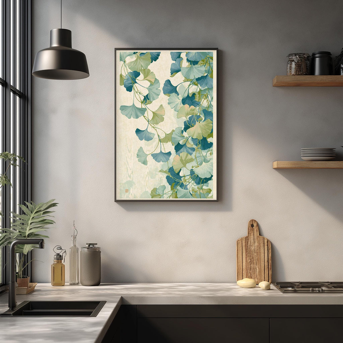 a kitchen counter with a framed print of green and white ginkgo leaves hanging above it. The counter also has various kitchen items such as a cutting board, a soap dispenser, and a bottle.