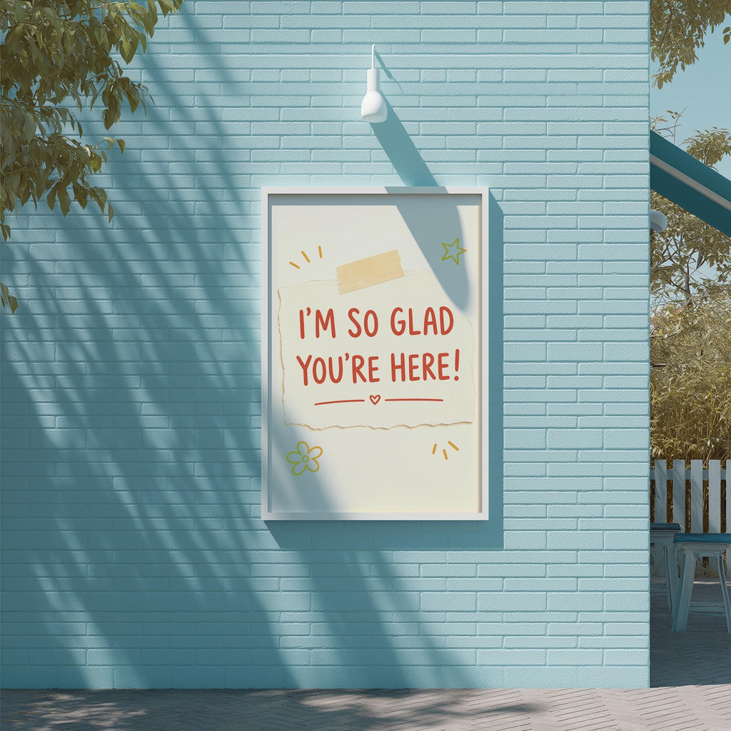 A framed poster with the text "I'm so Glad You're Here!" displayed on a blue brick wall.