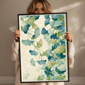 A woman holds up a framed print with a vibrant, leafy pattern of green and blue ginkgo leaves.