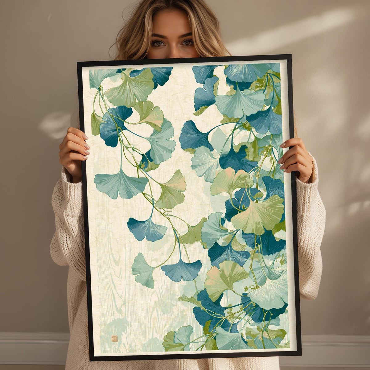 A woman holds up a framed print with a vibrant, leafy pattern of green and blue ginkgo leaves.