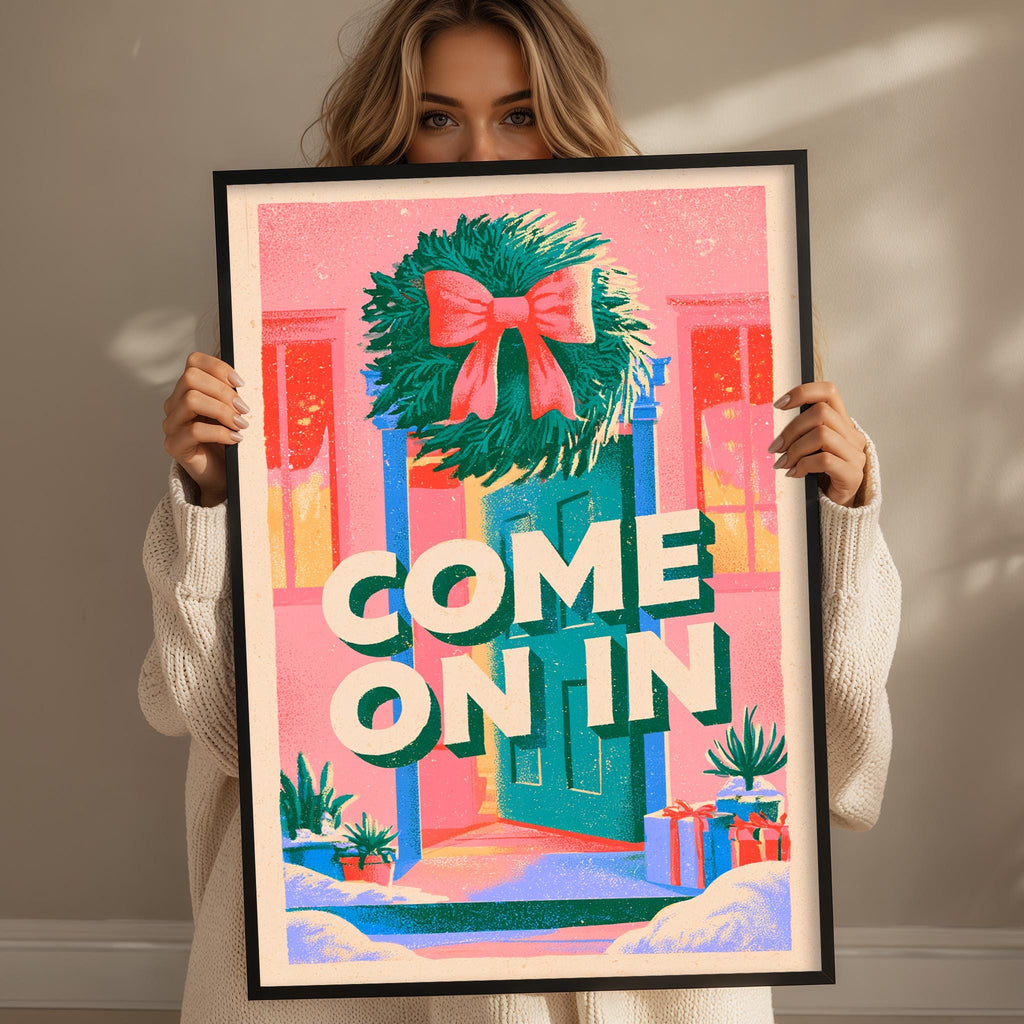 A woman holding a framed poster with a colorful illustration of a wreath and the text "COME ON IN"