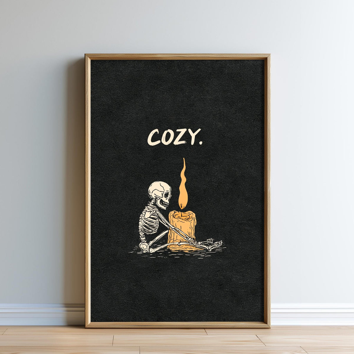 A framed poster with a skeleton sitting next to a lit candle, with the word "COZY" written above the image.