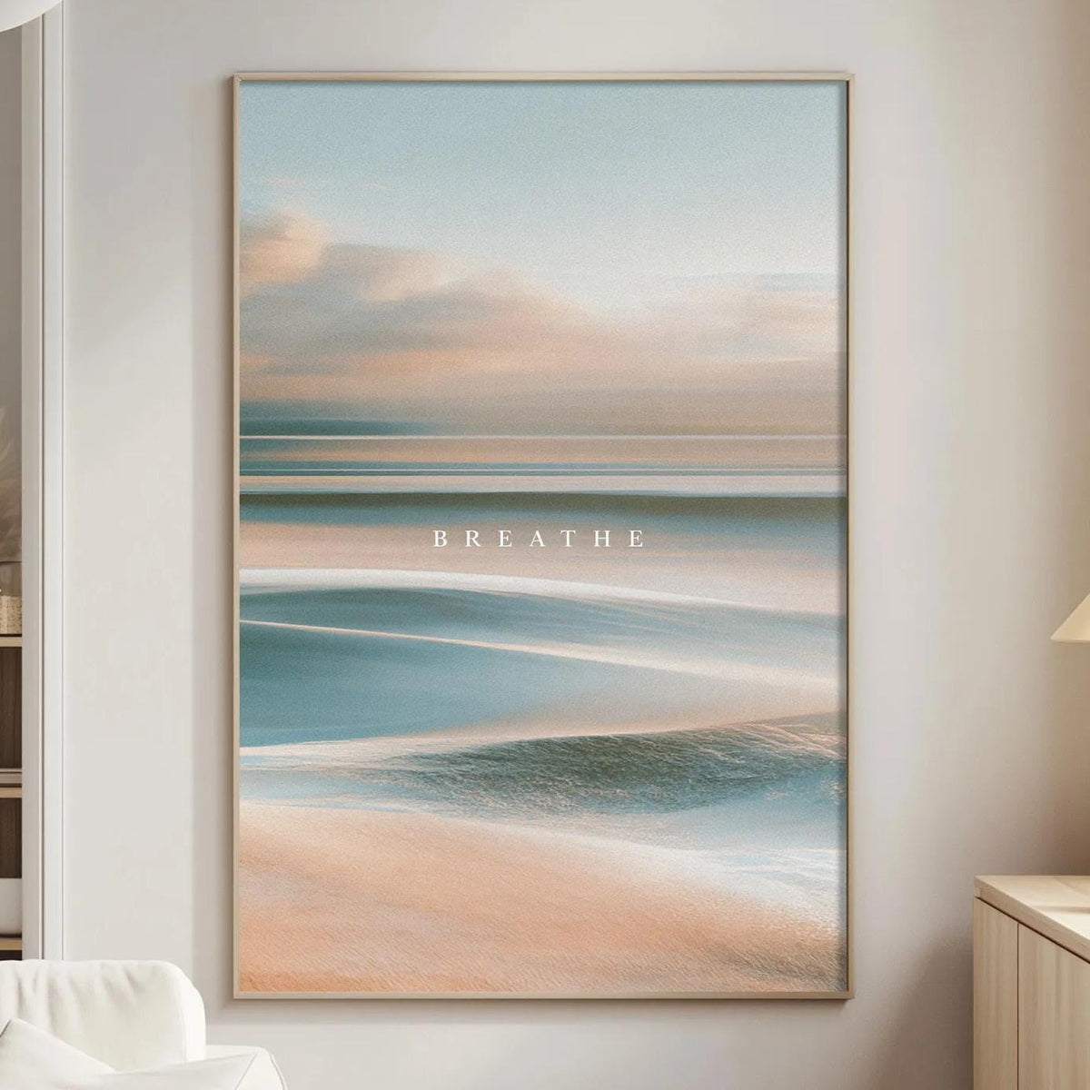 a serene living room with a large framed picture of a beach scene hanging on the wall. The picture captures the ocean and sky with a warm, soothing color palette. The room is furnished with a white sofa and a wooden side table, creating a cozy and inviting atmosphere.