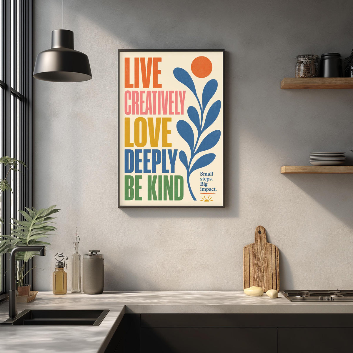 a kitchen counter with a framed poster hanging on the wall above it. The poster has a colorful design with text that reads "Live Creatively, Love Deeply, Be Kind" and includes a small sun symbol. The kitchen counter has various items on it, such as a cutting board, a soap dispenser, and a bottle.