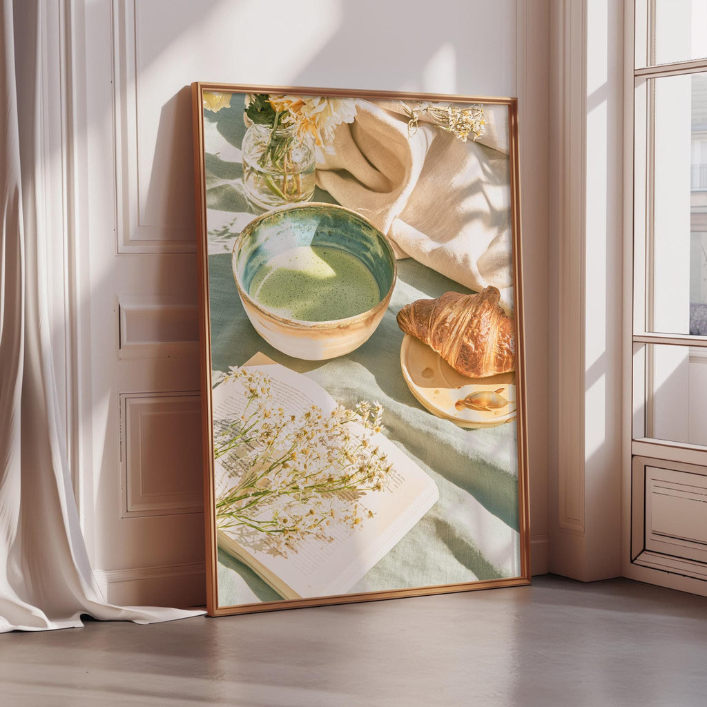 A framed print depicting a cozy breakfast scene with a croissant, a bowl of soup, and a vase of flowers on a table.