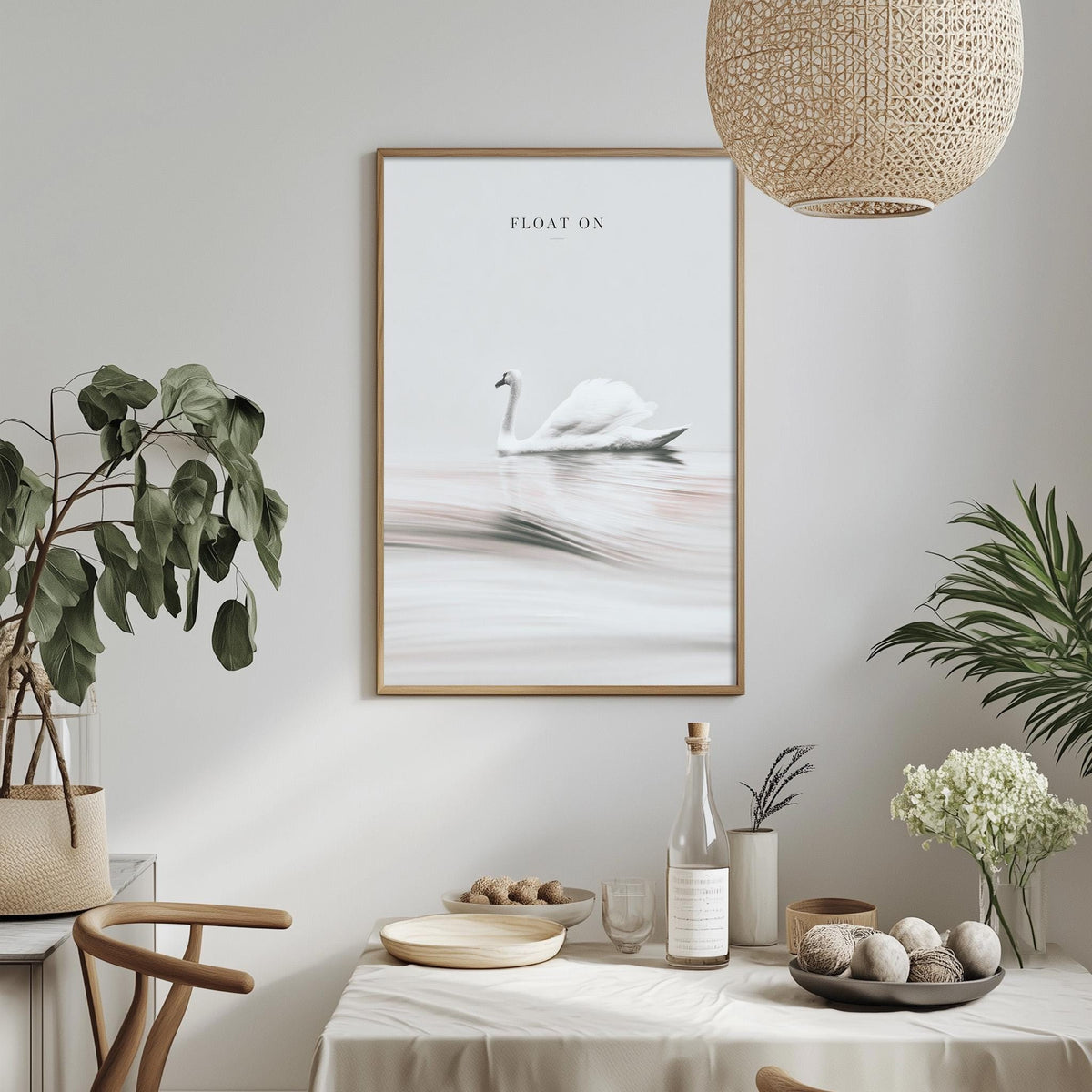 a dining table with a white tablecloth, a bowl of fruit, a bottle, and a vase with flowers. Above the table, there is a framed picture of a swan floating on a lake.