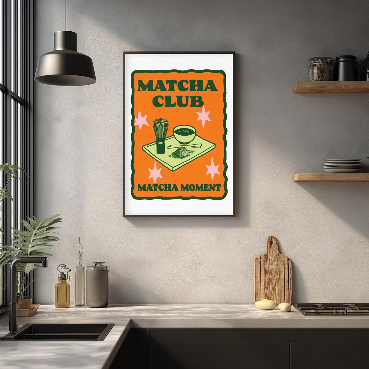 a framed poster with the text "MATCHA CLUB MATCHA MOMENT" and an illustration of a cup and a bowl on a table. The poster is hanging on a wall above a kitchen counter with various kitchen items such as a cutting board, a bottle, and a bowl.