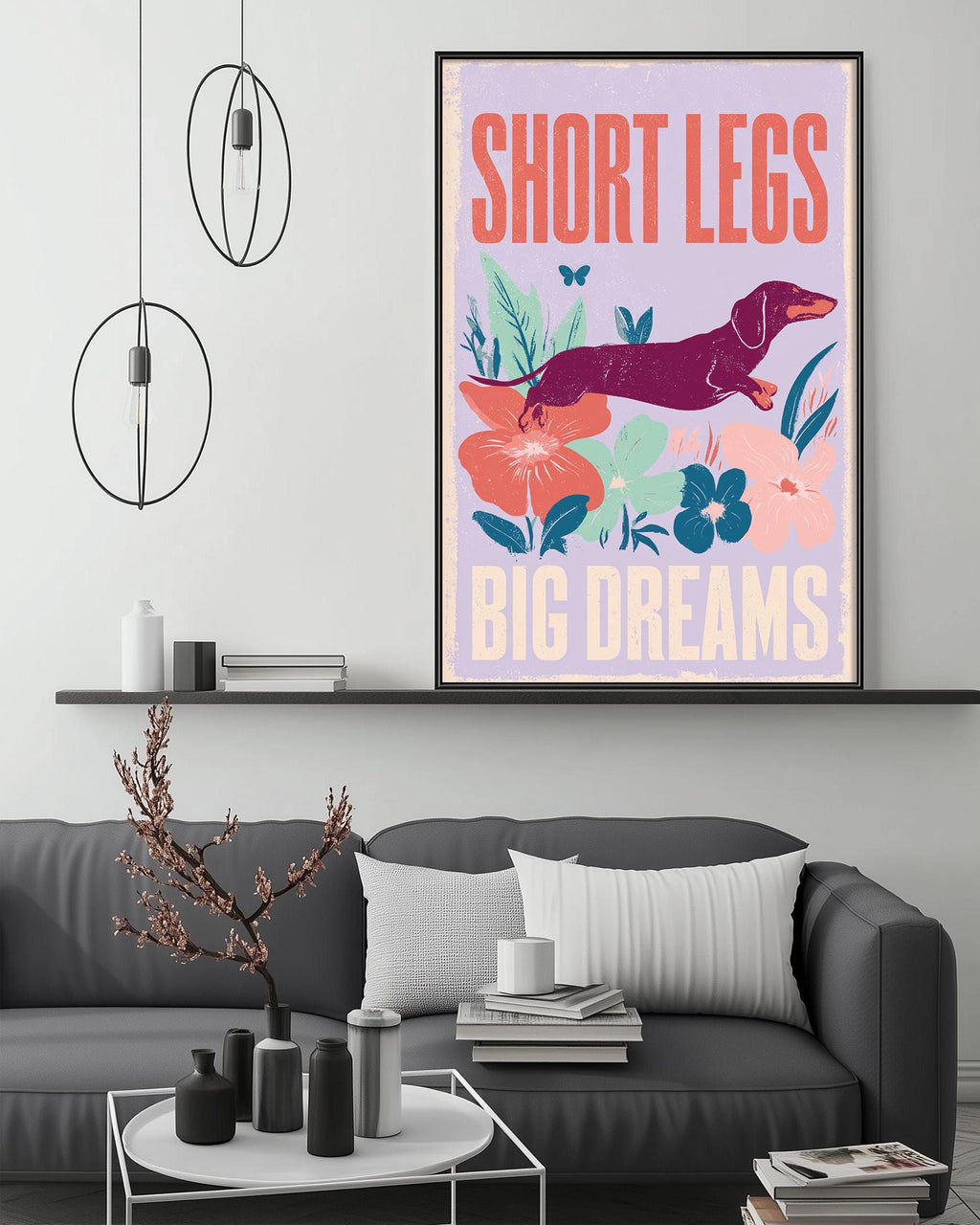 a poster with the text "SHORT LEGS BIG DREAMS" and an illustration of a dachshund dog. The poster is displayed on a wall above a gray couch in a living room setting.
