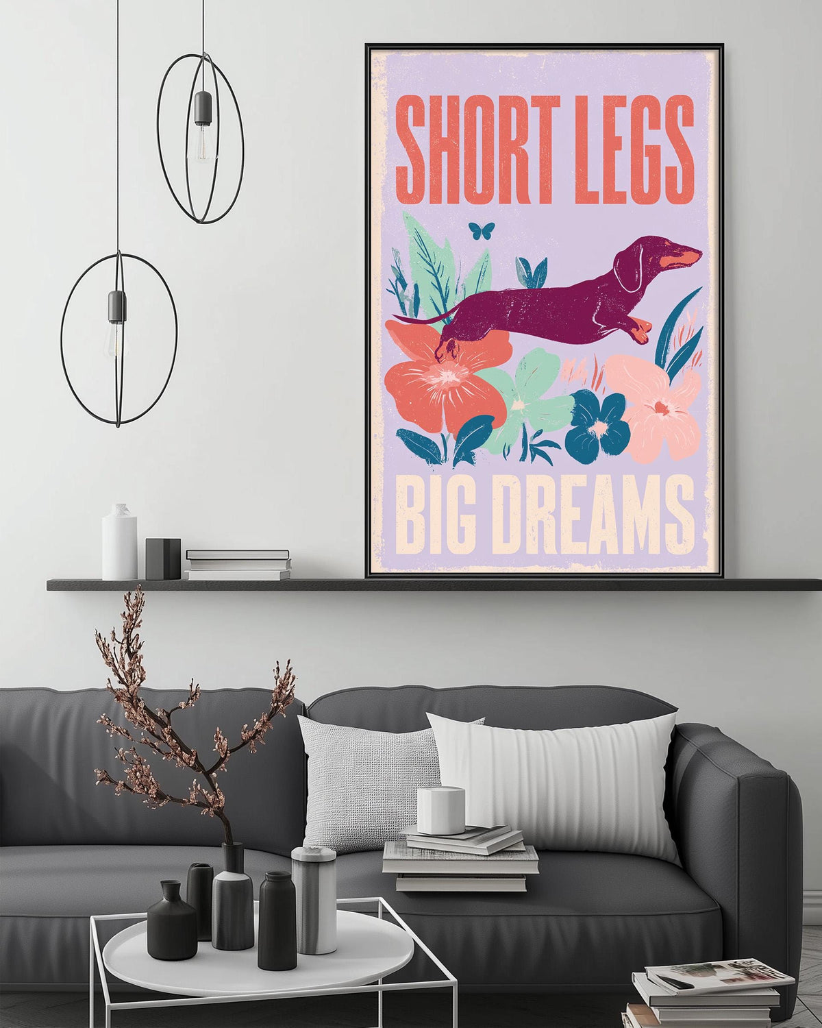 a poster with the text "SHORT LEGS BIG DREAMS" and an illustration of a dachshund dog. The poster is displayed on a wall above a gray couch in a living room setting.