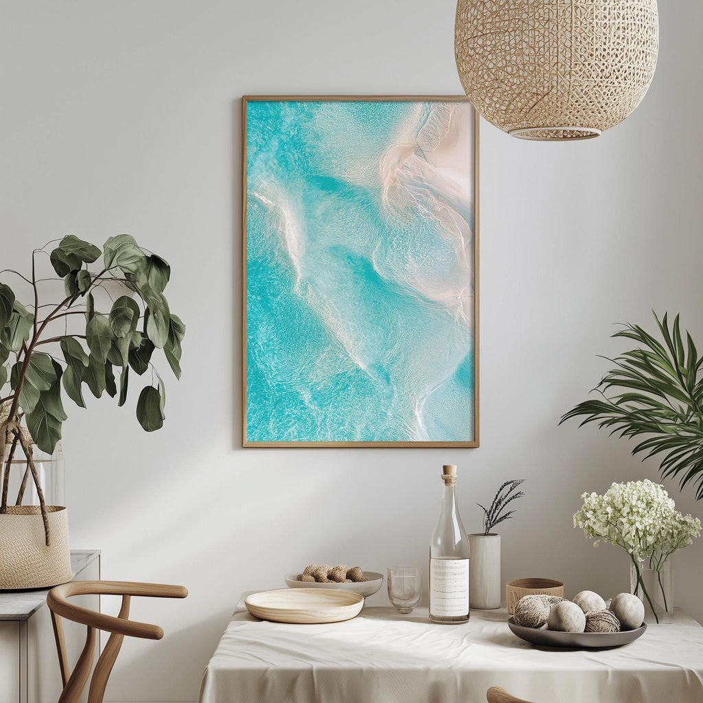 a dining table with a white tablecloth, a bowl of fruit, a bottle, and a vase with flowers. Above the table, there is a framed picture of a beach scene with turquoise water and white sand.