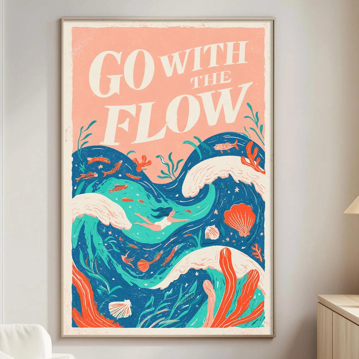a vibrant poster with the text "GO WITH THE FLOW" displayed prominently on it, set against a colorful ocean scene with waves, coral, and marine life. The poster is hanging on a wall in a living room, with a white couch and a small table nearby.