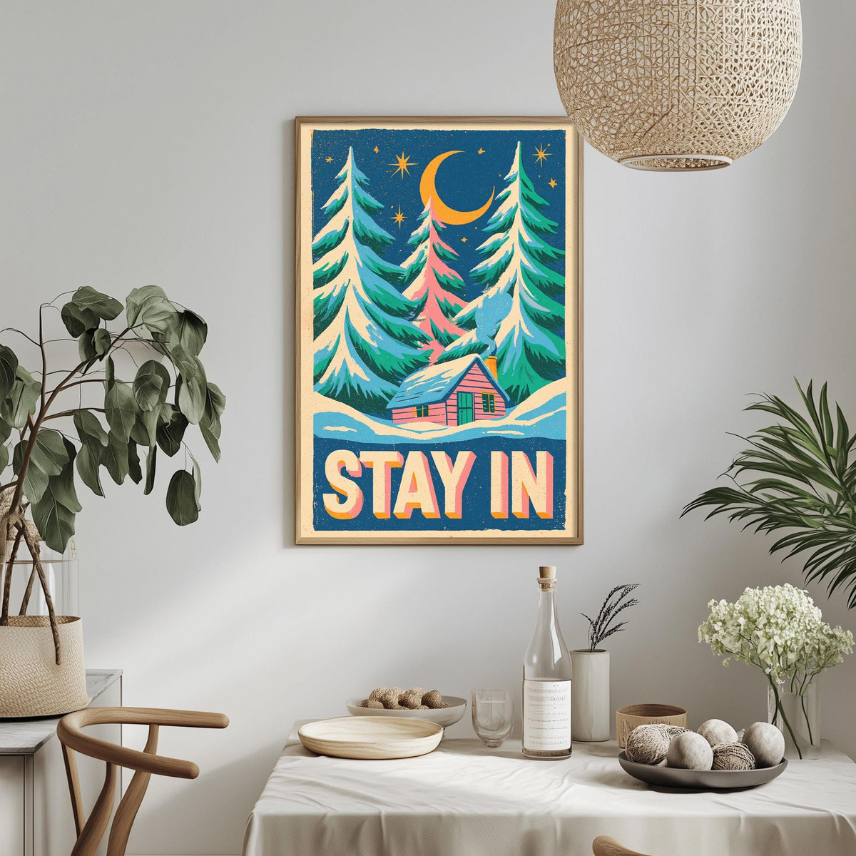a framed poster with a winter scene, including pine trees, a cabin, and a crescent moon, hanging on a wall above a dining table with various items on it.