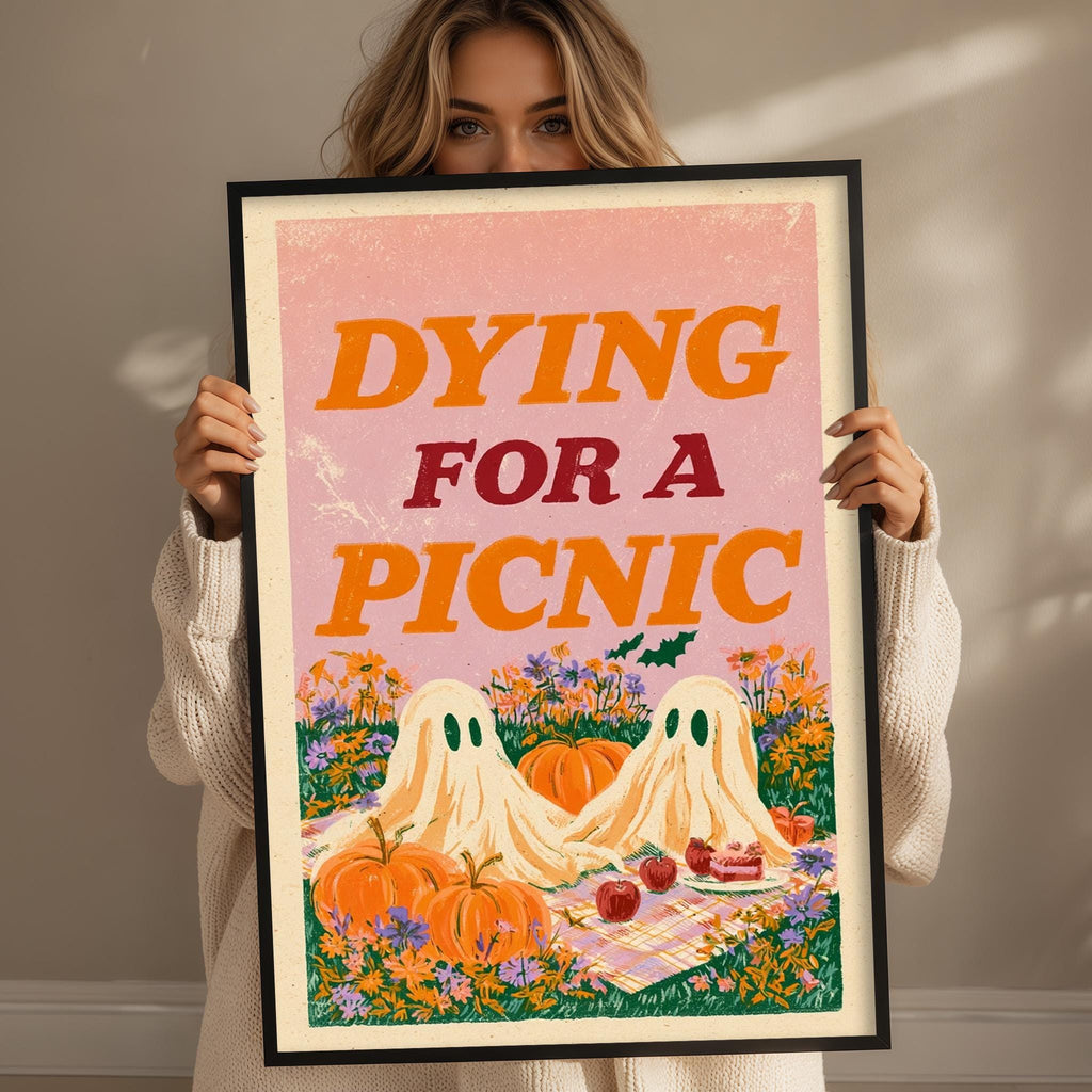 A woman is holding up a framed poster with a Halloween-themed illustration of two ghostly figures sitting on a picnic blanket, surrounded by pumpkins, flowers, and a cake.