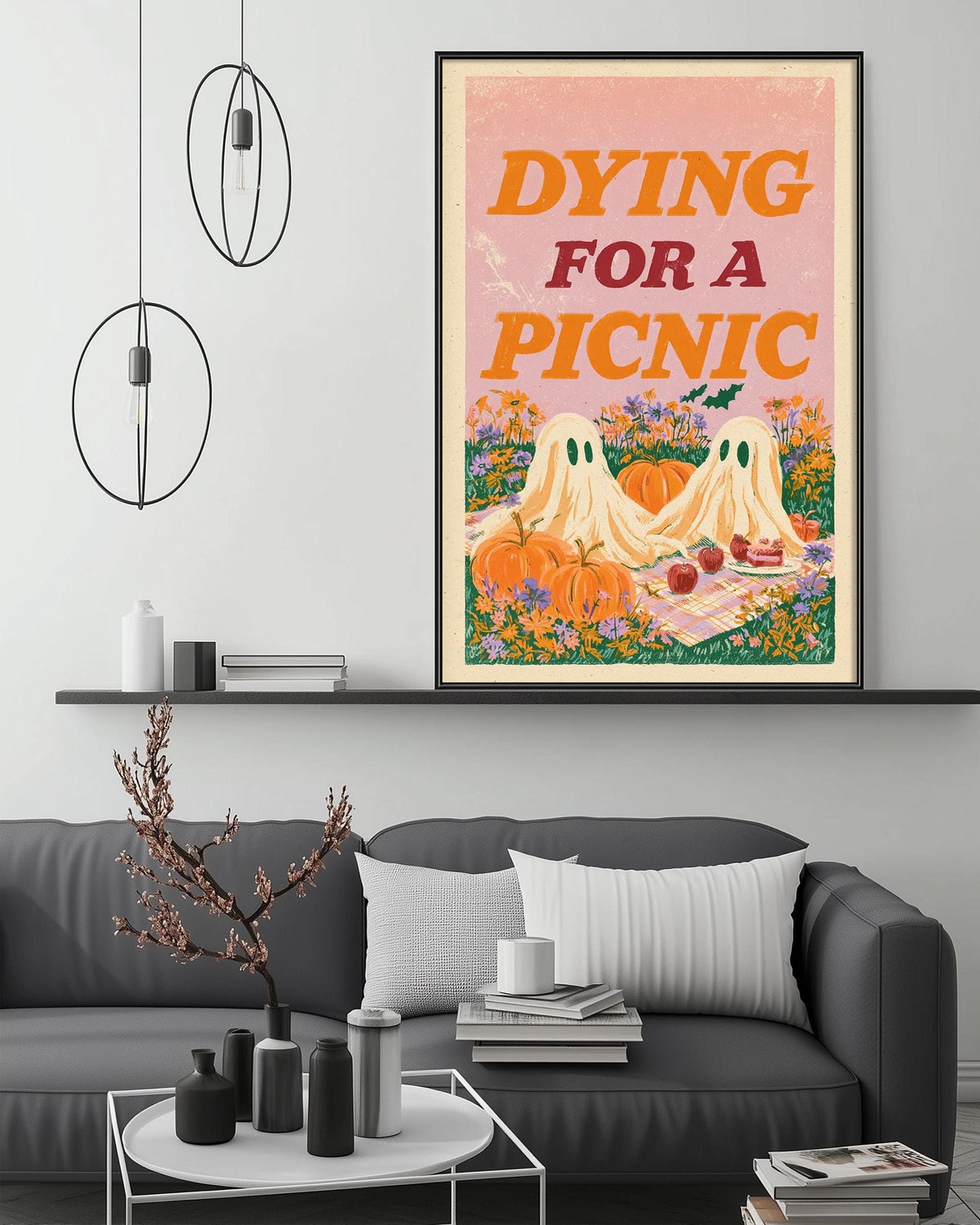 a living room with a gray couch, a coffee table, and a framed poster hanging on the wall. The poster depicts two ghostly figures and pumpkins, with the text " Dying for a Picnic" written on it.