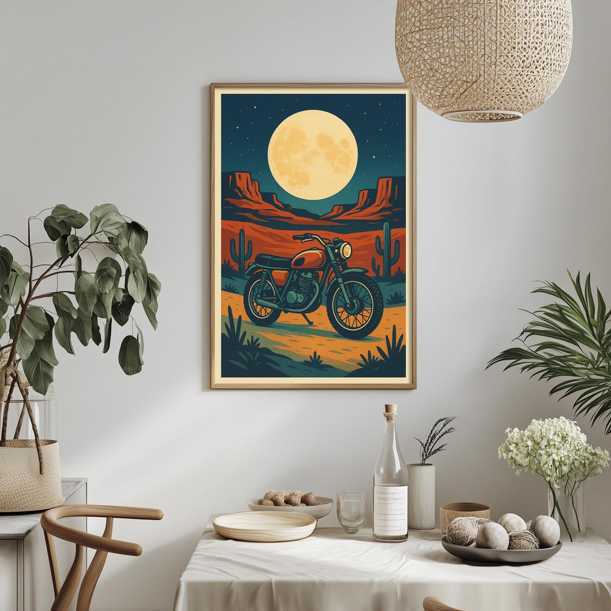 a framed poster of a motorcycle on a desert road, with a large full moon in the background. The poster is hanging on a wall, and there is a dining table with a vase of flowers, a bottle, and a bowl of fruit in the foreground.