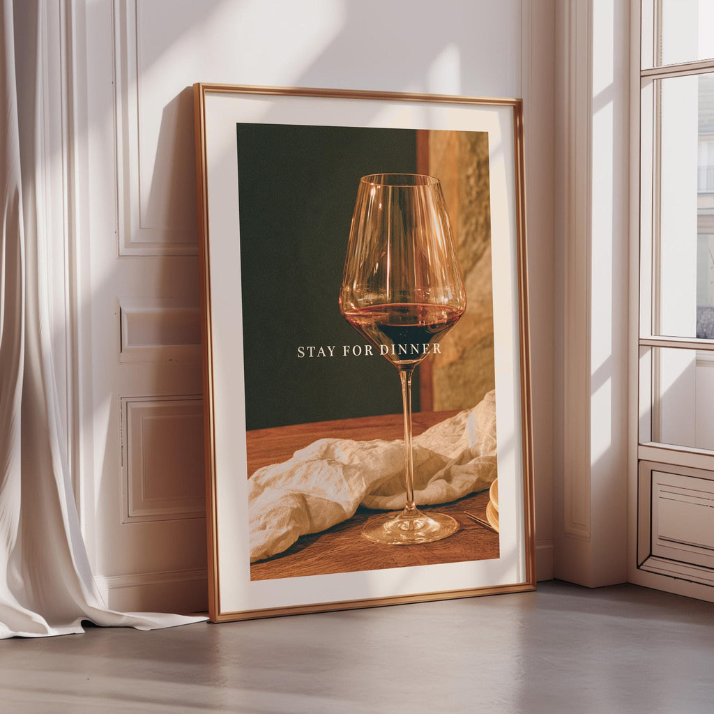 A framed poster with a wine glass and a napkin on a table, along with the text "STAY FOR DINNER" displayed on the poster.