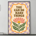 a poster with the text "YOU CAN DO HARD THINGS" displayed on a wall above a gray couch with a coffee table in front of it.