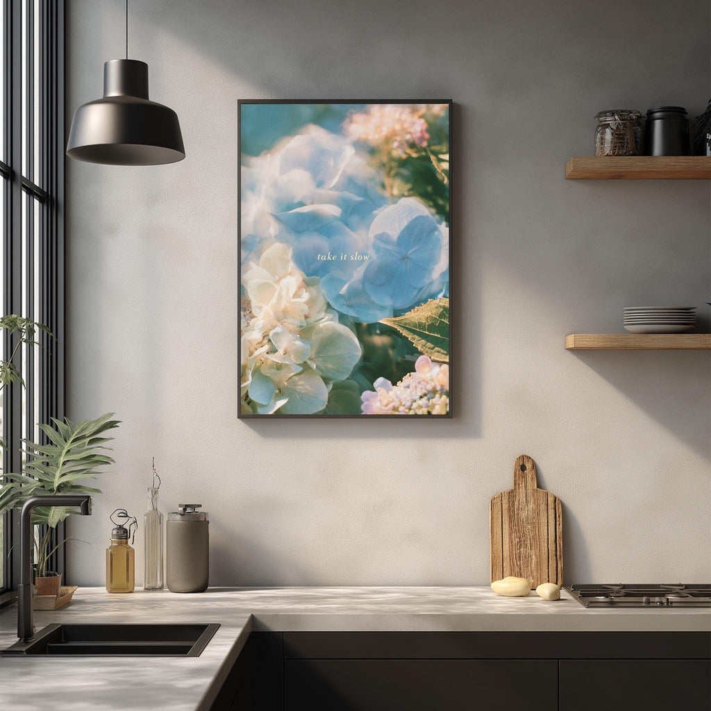 a kitchen counter with various items such as a cutting board, a soap dispenser, and a plant. Above the counter, there is a framed picture of flowers hanging on the wall.
