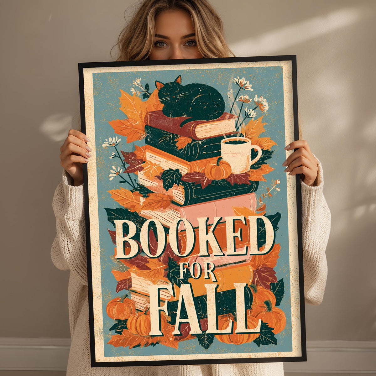 A woman holding a framed poster with a colorful autumn-themed design, including a stack of books, a cat, and flowers.