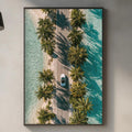 a kitchen counter with various kitchen items, including a cutting board, a bottle, and a vase. Above the counter, there is a framed picture of a tropical beach with palm trees and a car driving down a road.