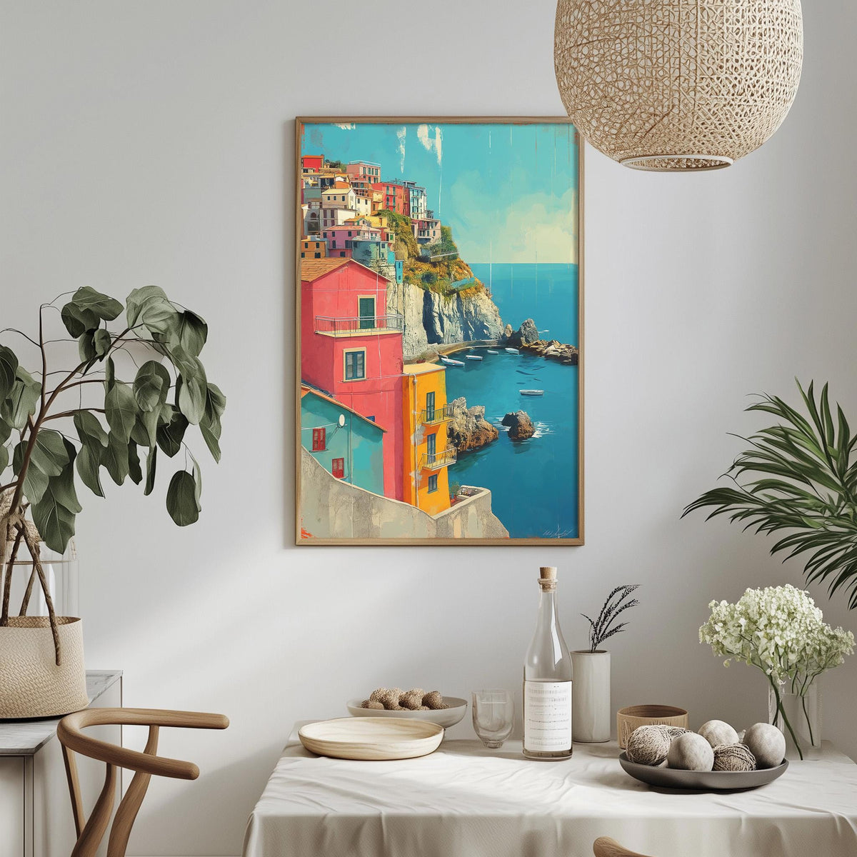 a dining table with a white tablecloth, a vase of white flowers, a bottle of wine, and a bowl of fruit. Above the table, there is a framed painting depicting a colorful coastal town with a blue sky and ocean in the background.