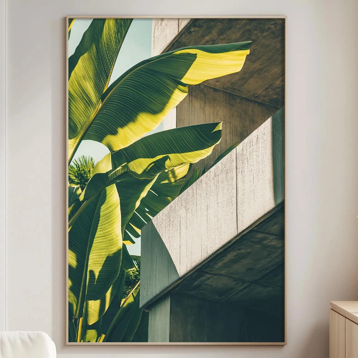 a large framed picture of a banana leaf on a wall, with a white sofa and a wooden side table in the foreground.