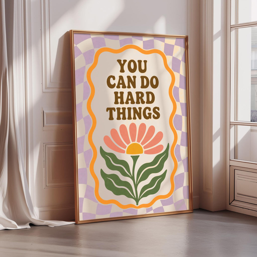 A framed poster with the text "YOU CAN DO HARD THINGS" and a flower graphic, placed on a wooden floor next to a window.