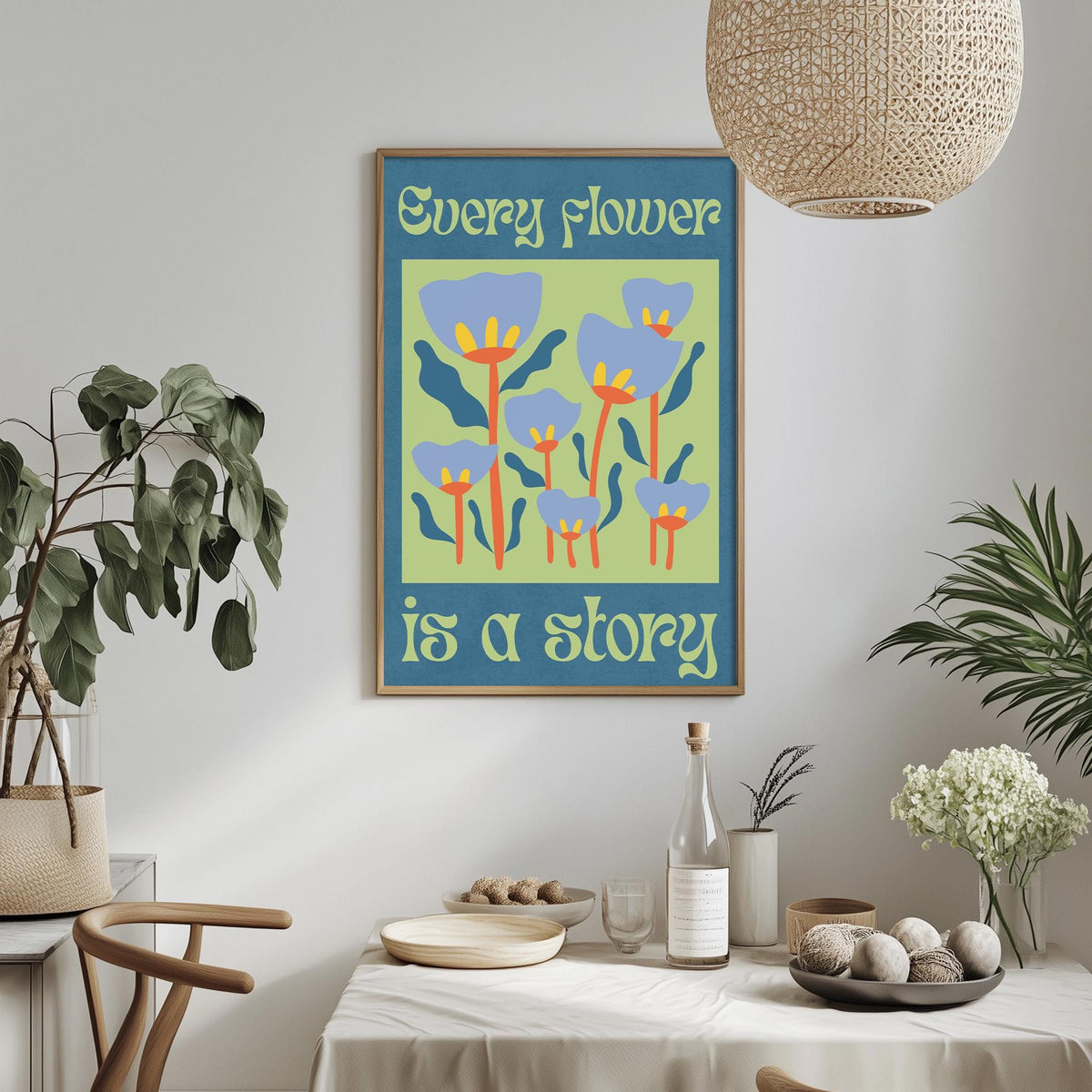 a dining table with a white tablecloth, surrounded by chairs. On the table, there are various items such as a bottle, a bowl, and a plate. Above the table, there is a framed poster with a colorful floral design and the text "Every flower is a story".