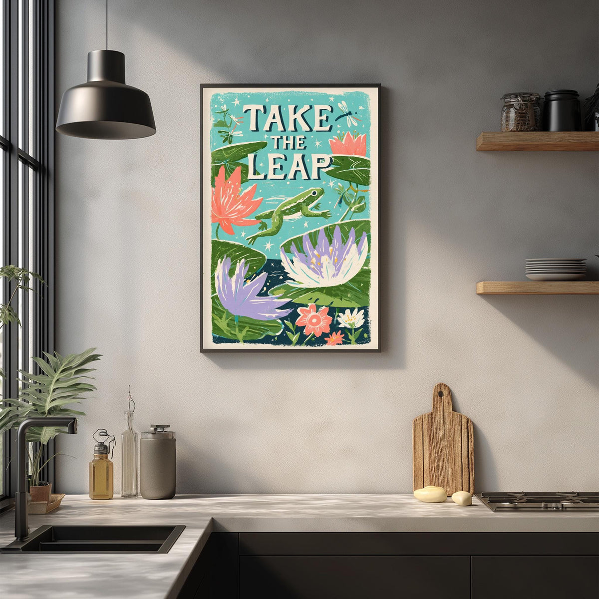 a kitchen counter with a framed poster of a frog leaping over a pond, surrounded by flowers and plants. The poster has the text "TAKE THE LEAP" written on it.