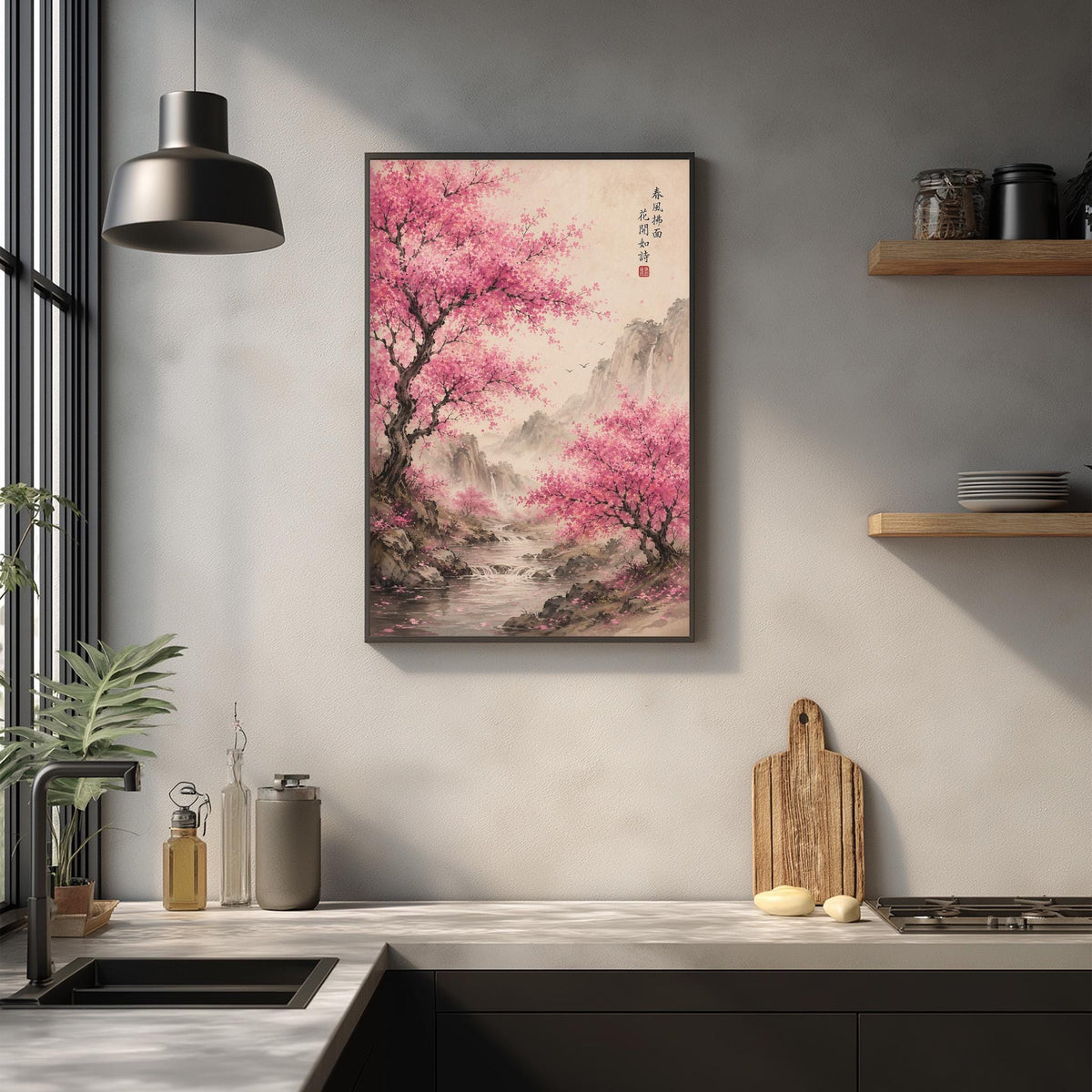 a kitchen counter with a framed picture of a pink cherry blossom tree hanging above it. The kitchen counter also has various kitchen items such as a cutting board, a bottle, and a vase.