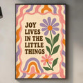 a kitchen counter with various items such as a cutting board, a soap dispenser, and a potted plant. Above the counter, there is a framed poster with the text "Joy lives in the little things" and a colorful flower design.